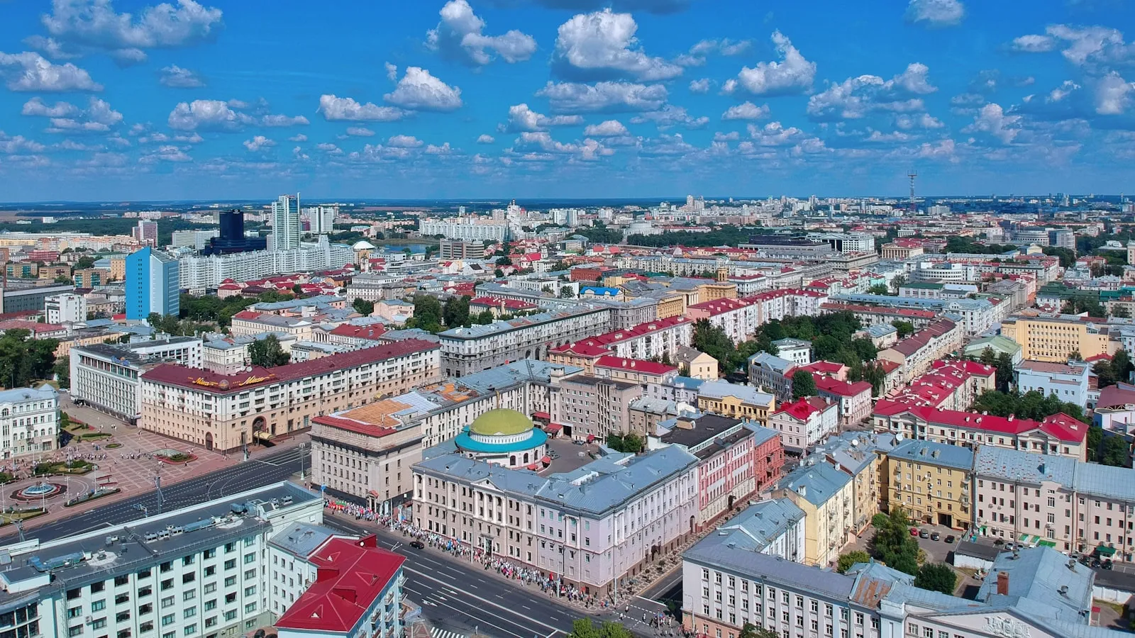 Aerial view of central Minsk with red-roofed blocks and wide avenues under a summer sky.
