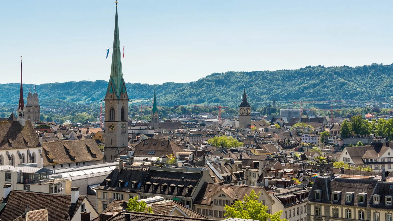 Rooftops and church spires across central Zurich, with wooded hills rising behind the city.