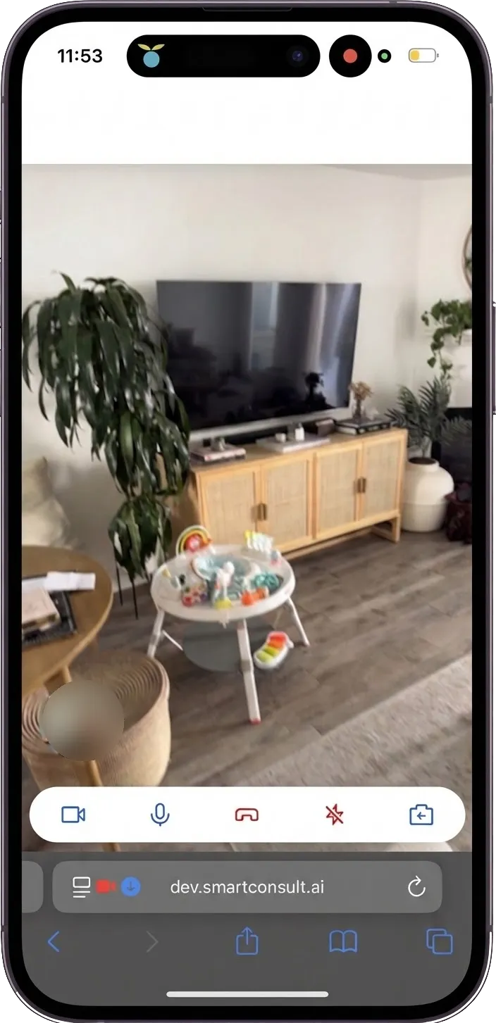 A phone on a live Yembo video call, showing a living room with a television, a sideboard, and plants, with call controls for camera, microphone, hang up, flash, and capture along the bottom.