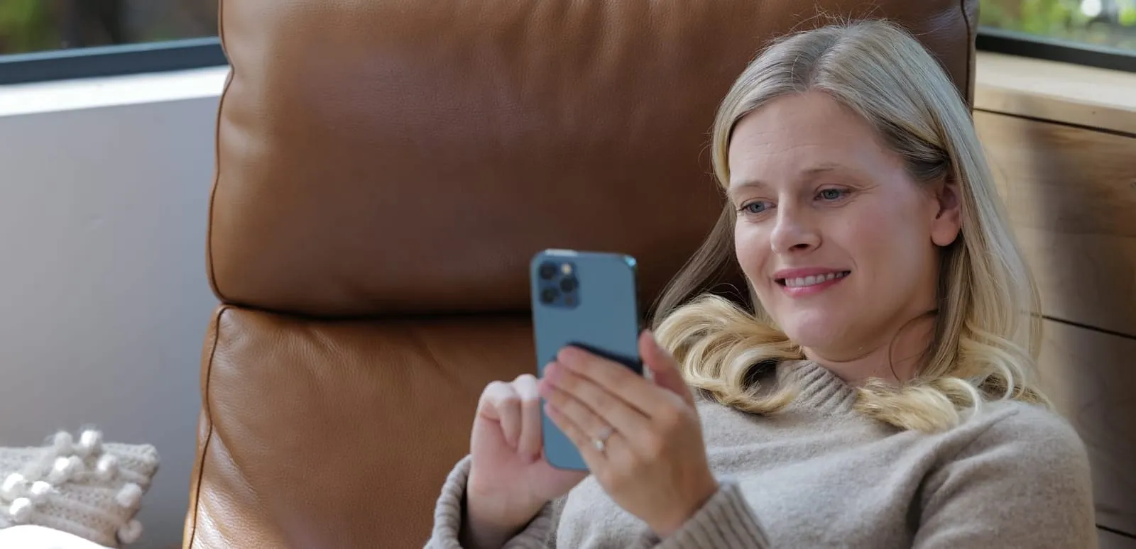 A woman sitting in an armchair by a window at home, reading on her phone.