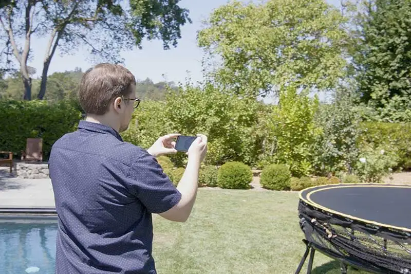 A homeowner standing in a backyard, photographing the garden and pool area with a phone.