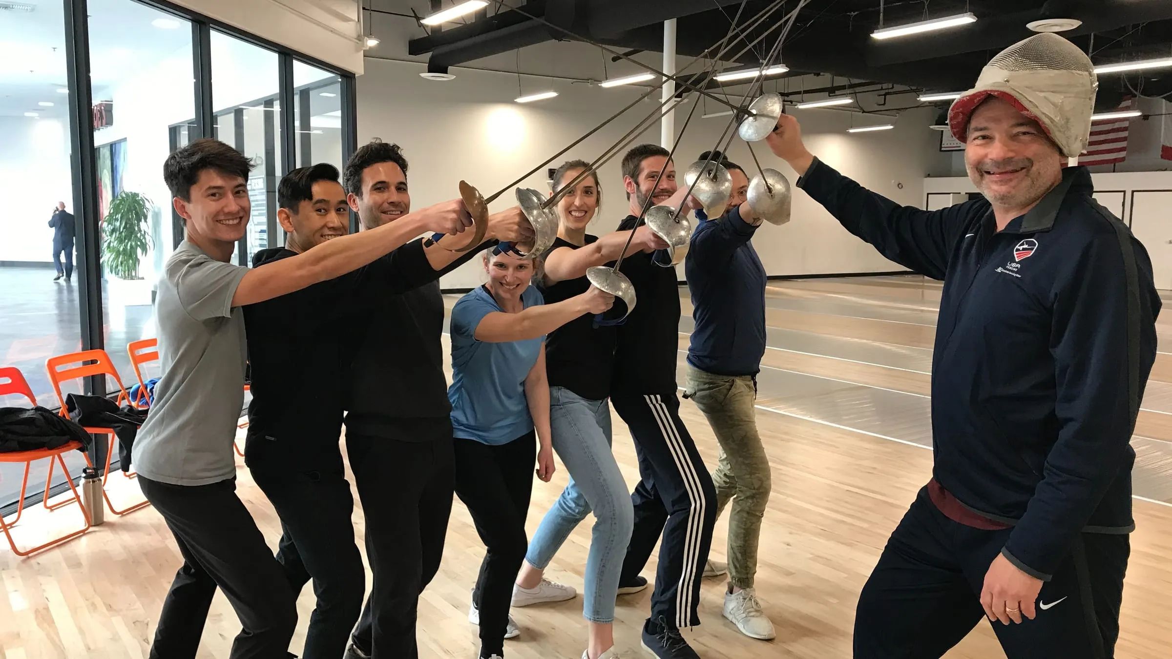 Eight members of the team on a fencing piste at a team outing, foils raised and crossed above them, with their instructor alongside.