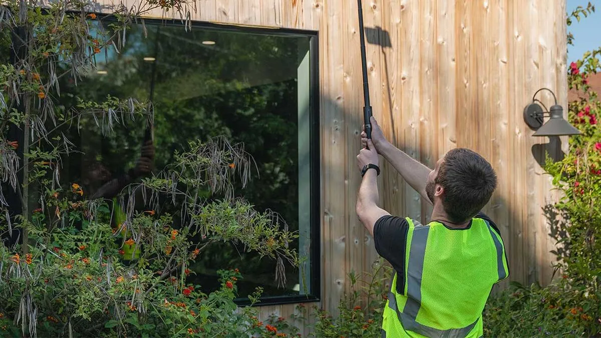An inspector in a high-visibility vest reaching up the side of a house with a pole-mounted camera.