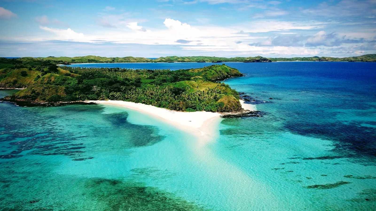 An aerial view of a white sand spit and turquoise lagoon off a forested island in Fiji.