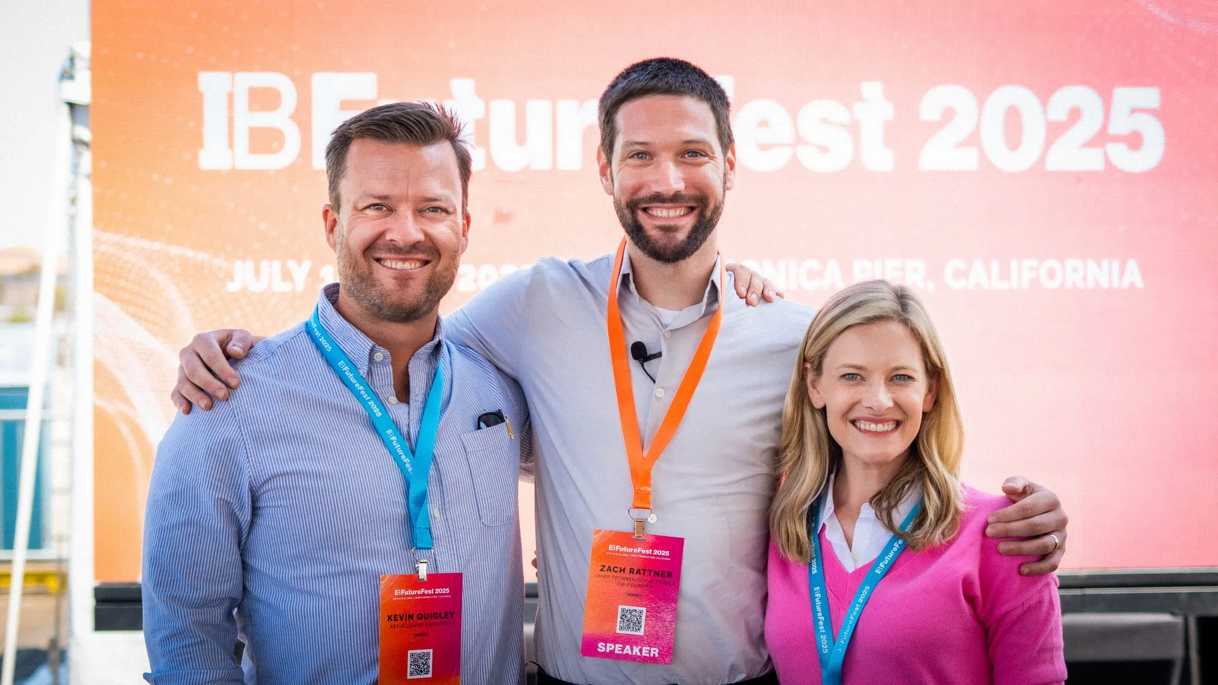 Three members of the Yembo team shoulder to shoulder in front of the IBFutureFest 2025 banner on the Santa Monica Pier, one of them wearing a speaker badge.
