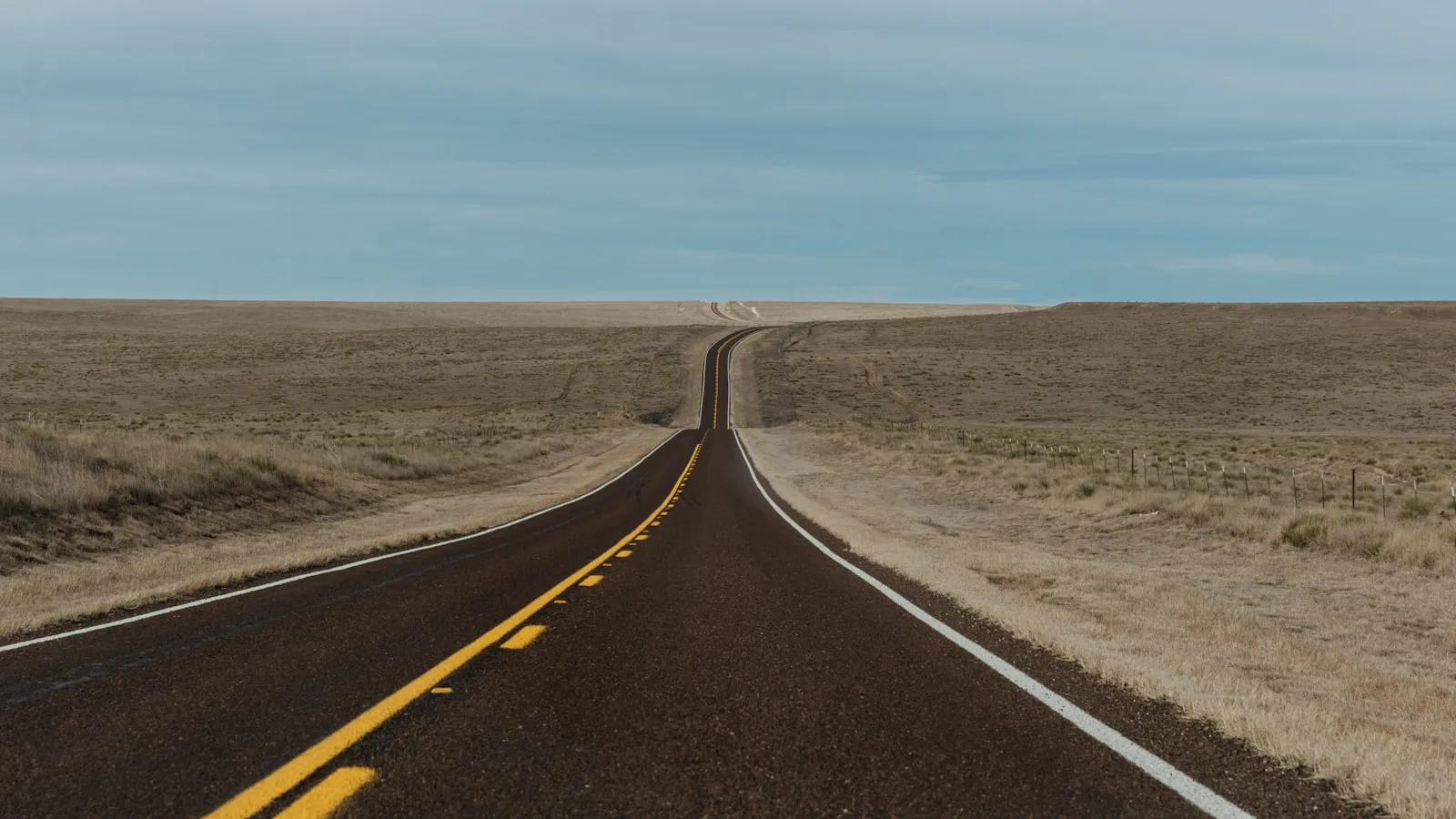 A long, empty road stretches through a dry, grassy landscape.
