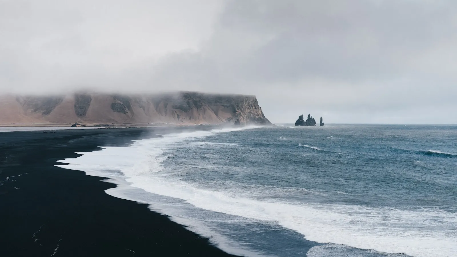 Waves crashing on a black sand beach below misty cliffs in Vik, Iceland.