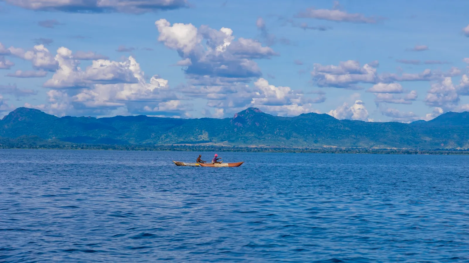 Two people kayak on a calm blue lake near mountains.