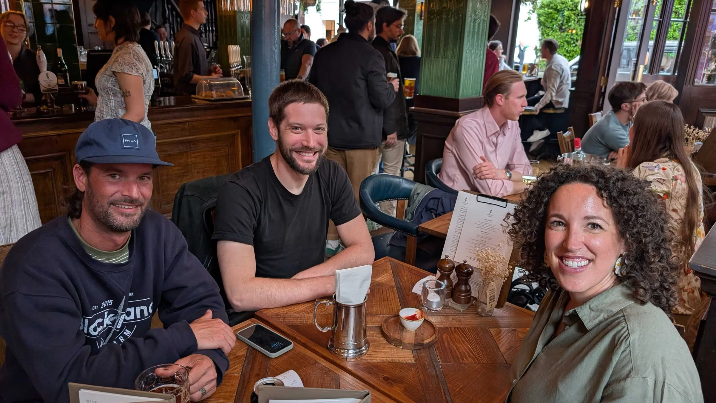 Three colleagues at a pub table on a work trip, smiling at the camera over drinks and menus in a busy room.