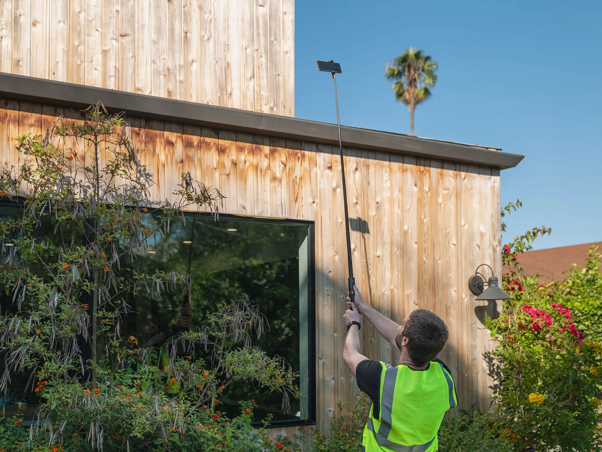An inspector in a high-visibility vest raises a phone on a telescoping pole to capture the roof of a wooden house.